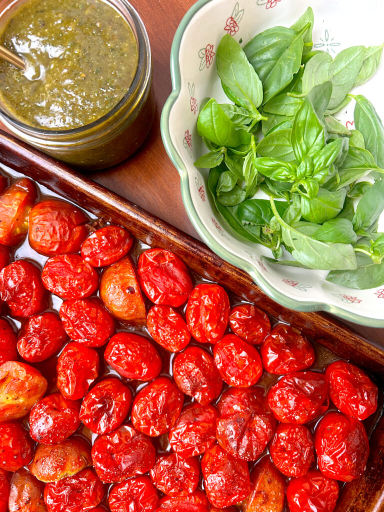 roasted tomatoes on a baking sheet with a jar of pesto and fresh basil