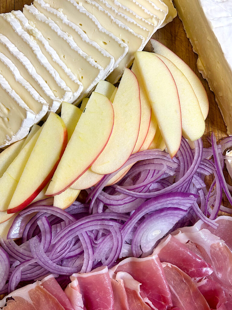 brie cheese, apples, red onion and prosciutto on a wood board