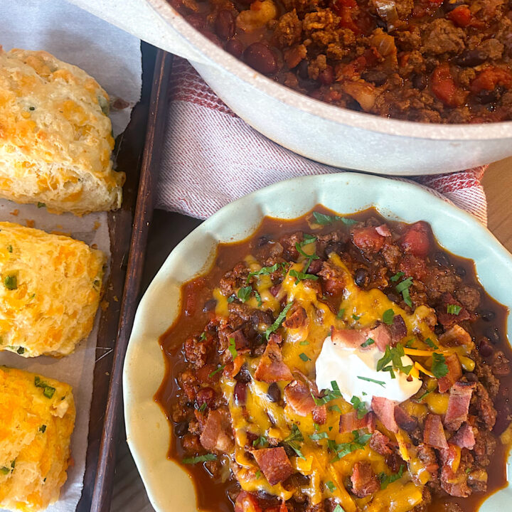 a green bowl full of venison chili with cheese, sour cream and cheddar biscuits