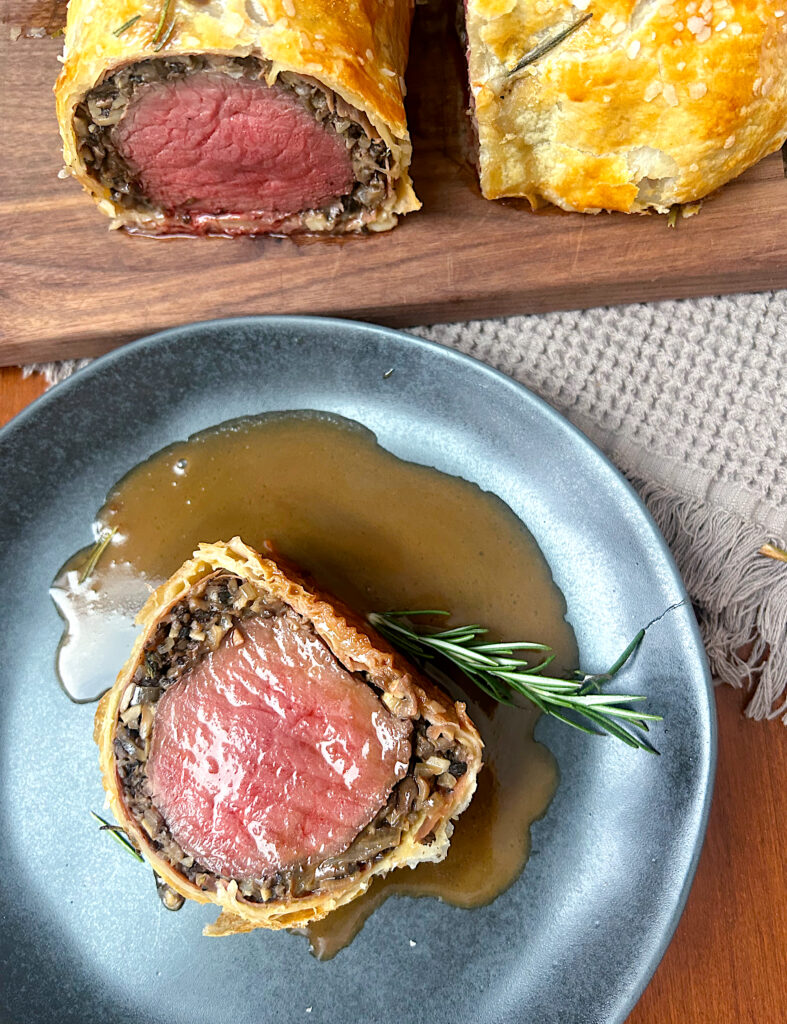 a black plate with a slice of tenderloin wrapped in mushrooms and puff pastry