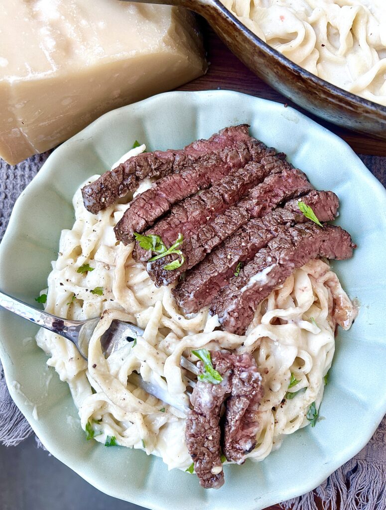 a green bowl of alfredo pasta with sliced steak
