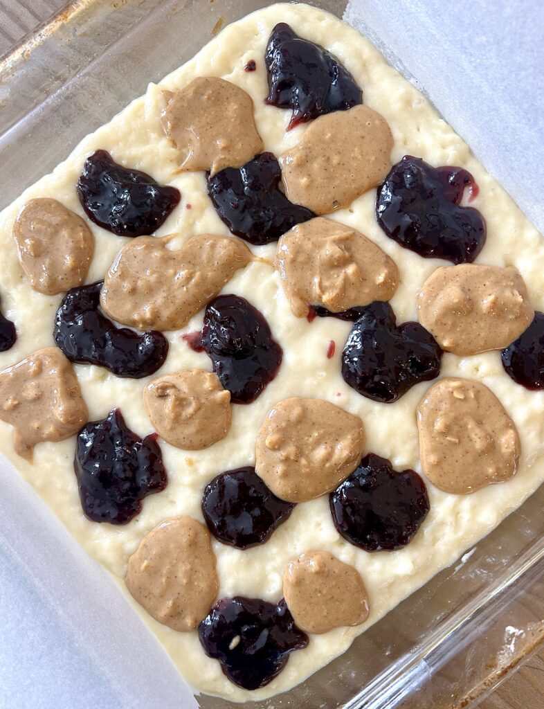 cake batter in a square pan with blobs of peanut butter and jam on top
