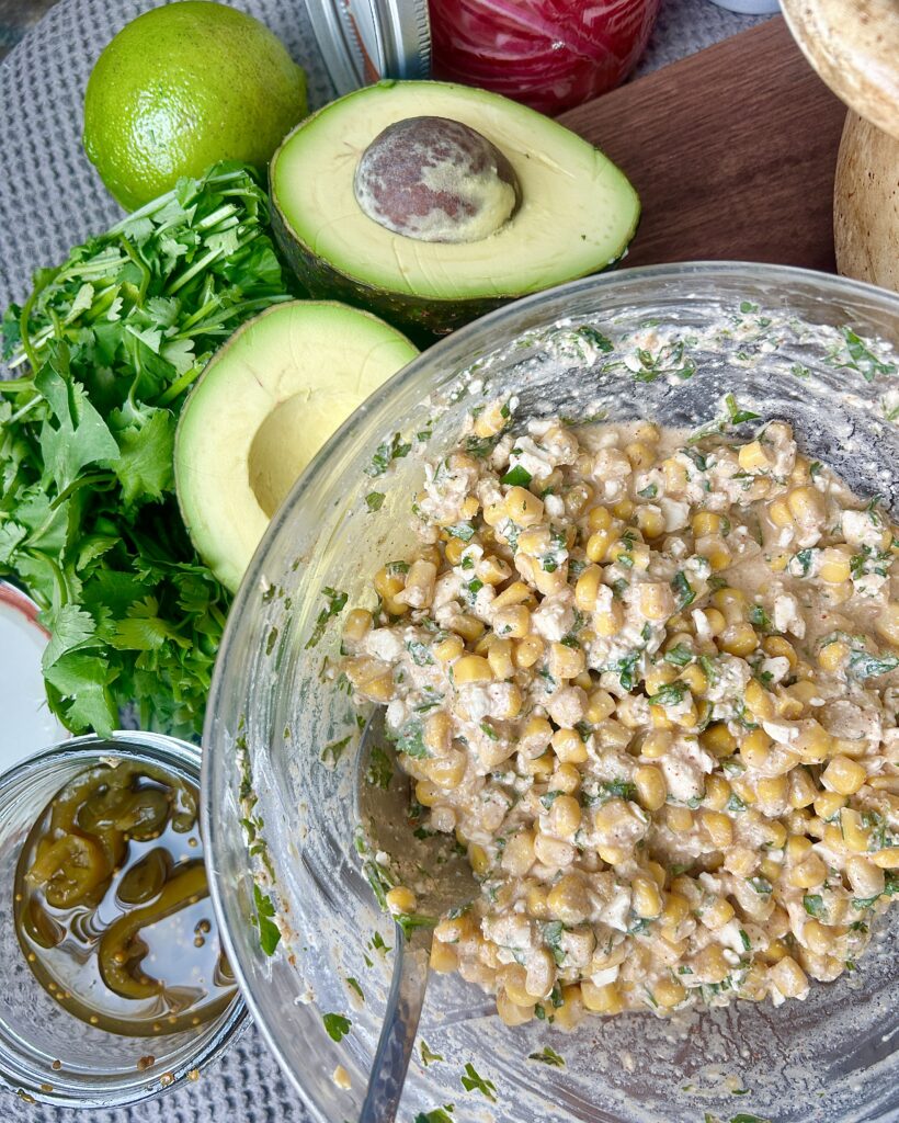 a bowl of street corn mixtures with avocados and cilantro on the side