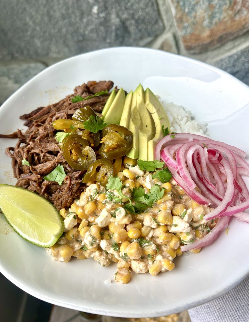 a white bowl with shredded meat, street corn, avocado and red onion