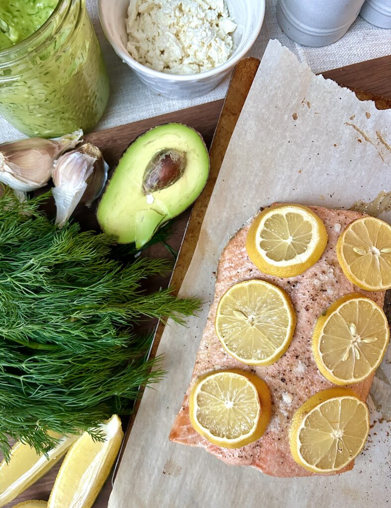 salmon with lemon slices on a baking sheet, with avocado and dill on the side