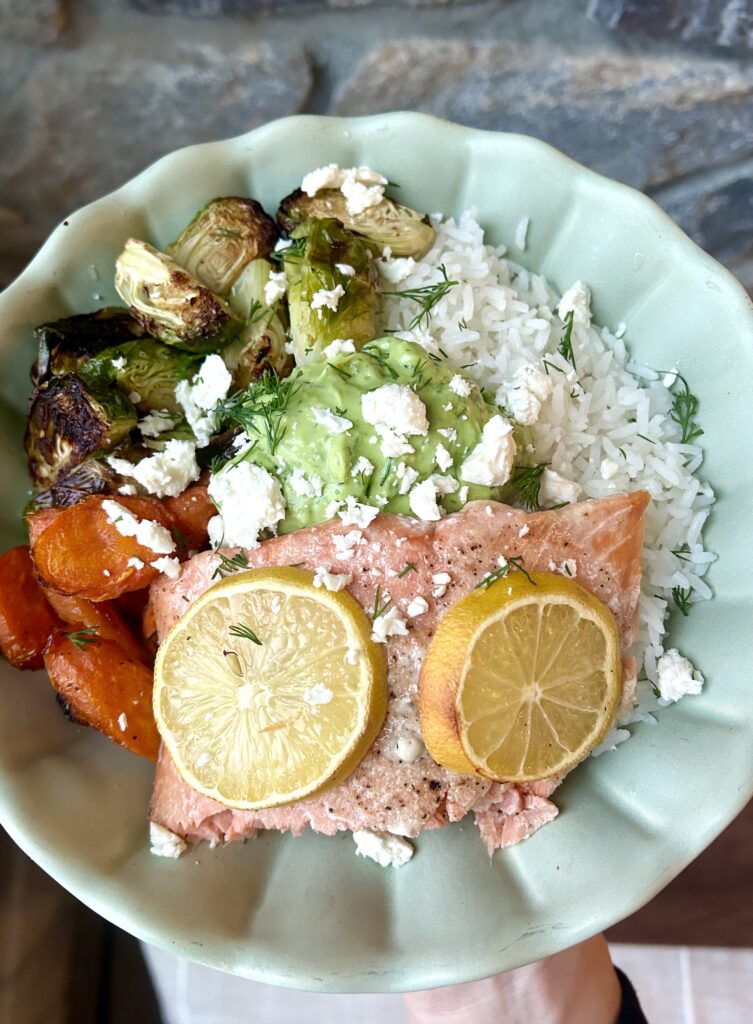 a green bowl with rice, roasted vegetables, salmon and a creamy avocado sauce
