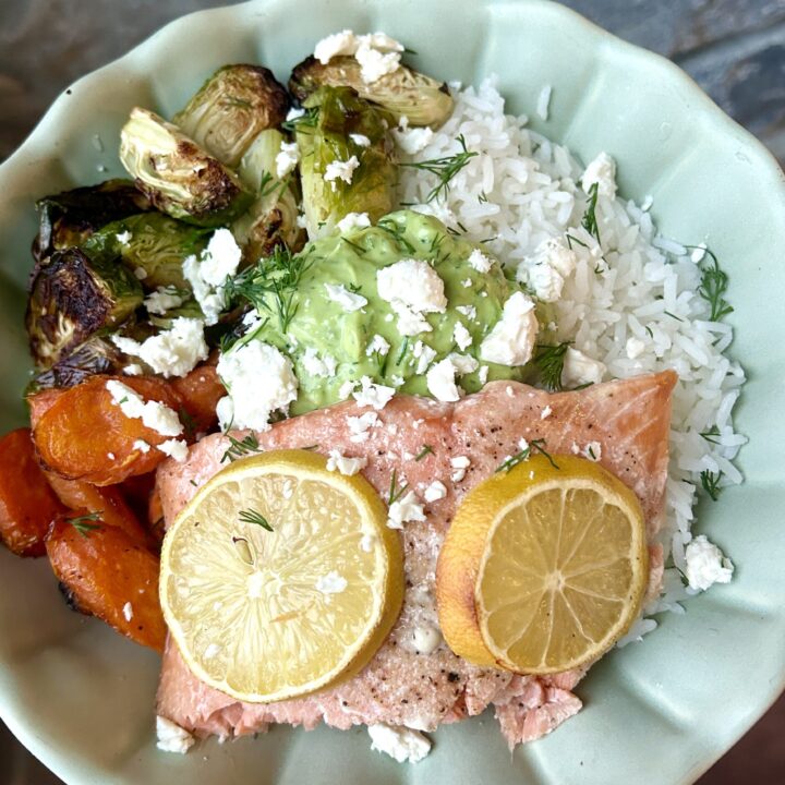 a green bowl with rice, roasted vegetables, salmon and a creamy avocado sauce