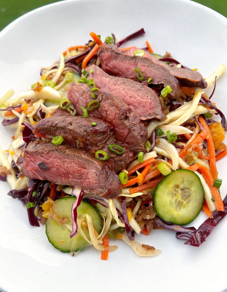 a bowl of crispy rice salad with carrots, cucumber, cabbage and sliced steak on top
