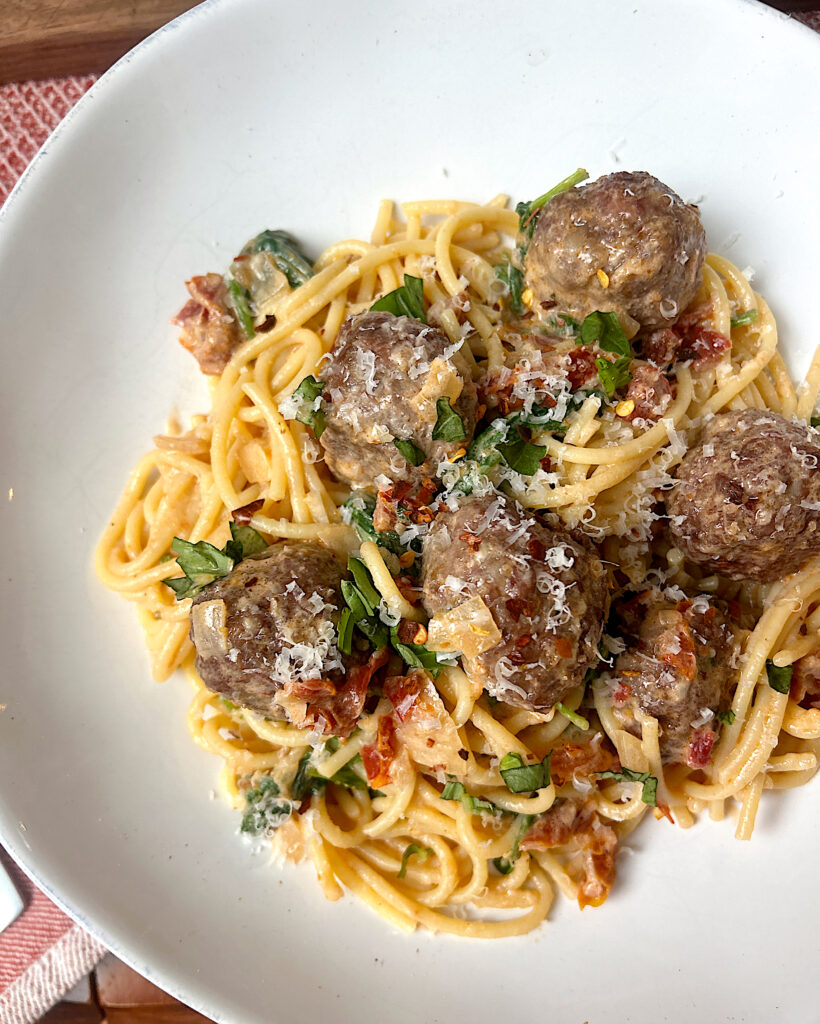 a white bowl of pasta with tomatoes, basil and meatballs