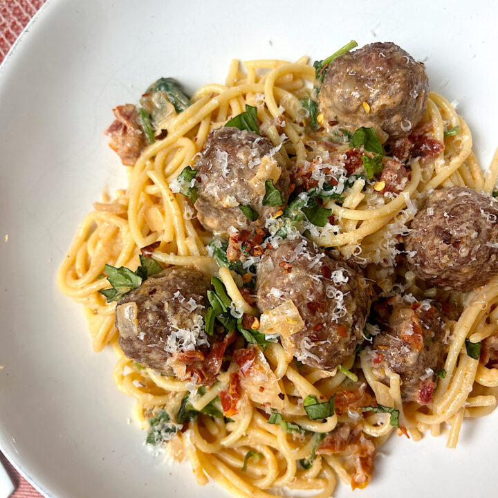 a white bowl of pasta with tomatoes, basil and meatballs