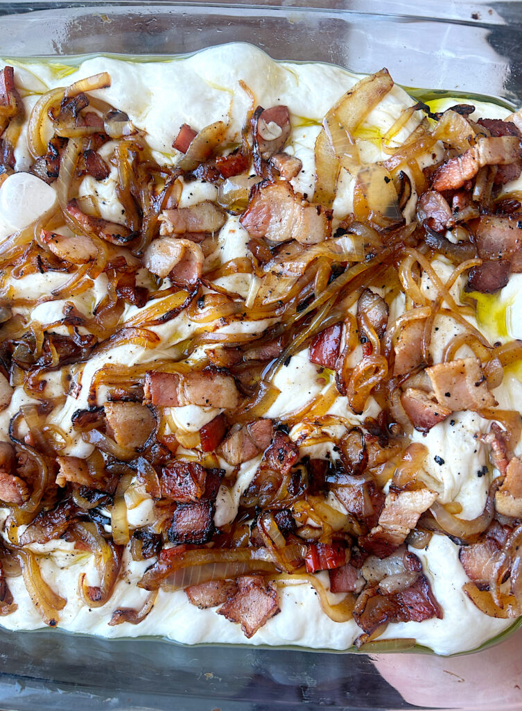 unbaked focaccia bread in a pan with bacon and onions on top