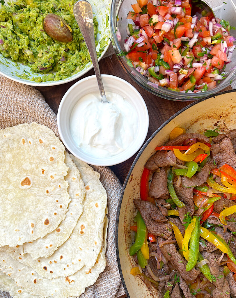 fajita peppers and steak in a pan with a side of tortillas, guacamole and sour cream
