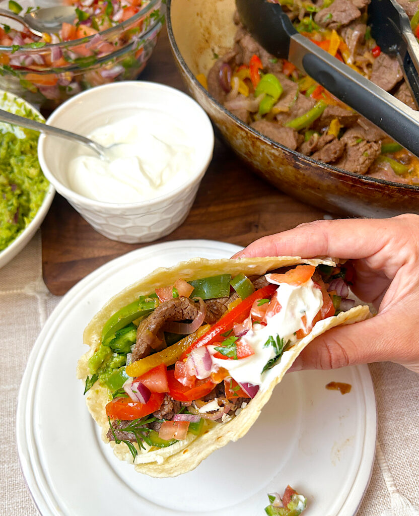 a white plate with a fajita full of venison steak , peppers, onions, salsa and sour cream
