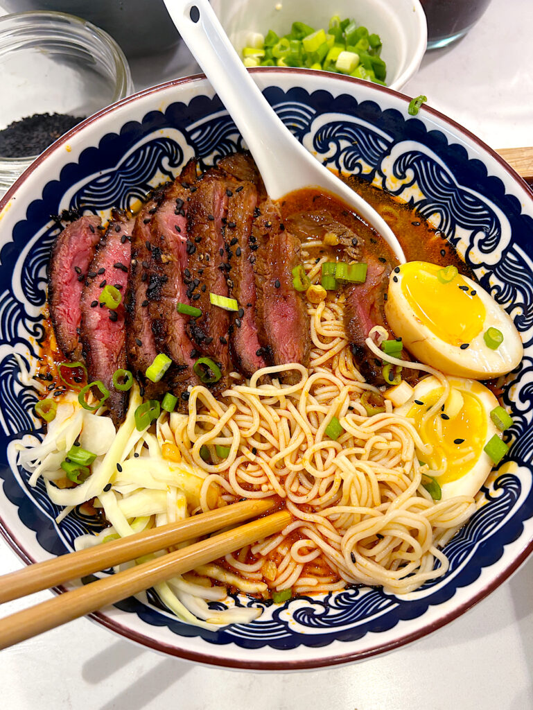 a blue bowl of Ramen noodle soup with steak and a soft boiled egg