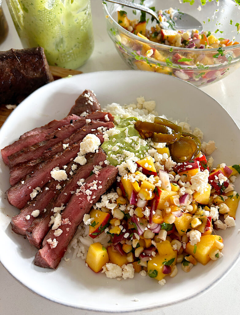 a white bowl of rice with slice steak, peach and corn salsa, and avocado sauce