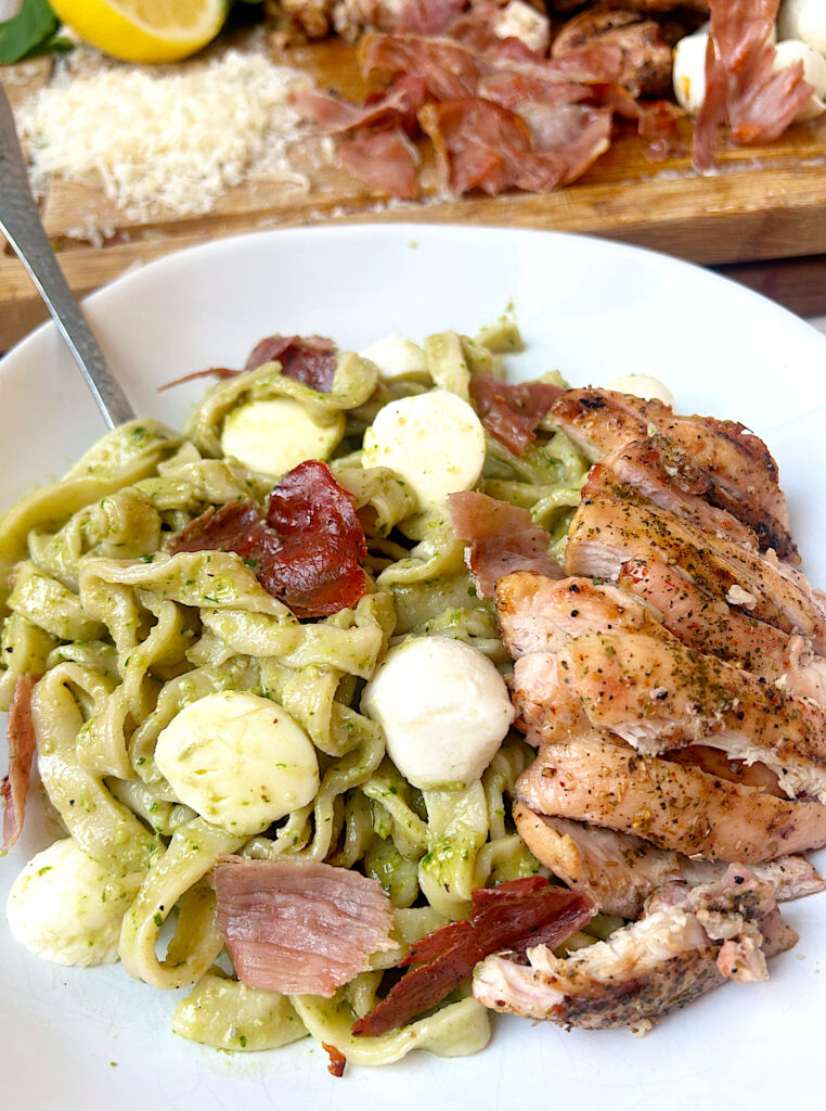 a white bowl of pasta with pesto sauce, chicken, prosciutto and mozzarella cheese