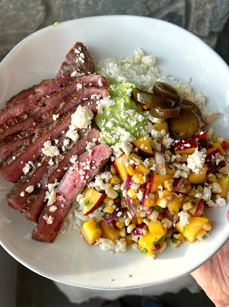 a white bowl with rice, sliced steak, peach and corn salsa and avocado sauce