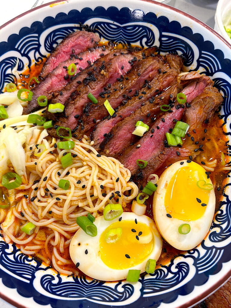 a blue bowl with Ramen noodles, chili broth, steak, and a boiled egg