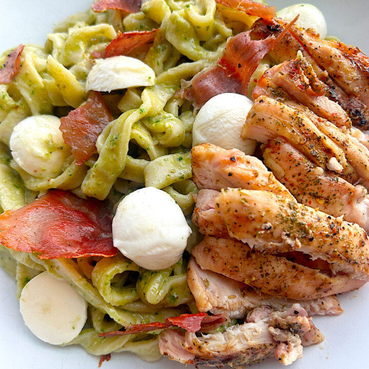 a white bowl of pesto pasta with prosciutto, chicken and mozzarella balls