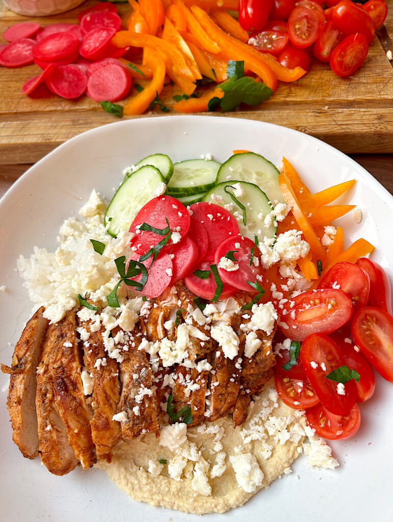 a white bowl with hummus, rice, vegetables and spiced chicken