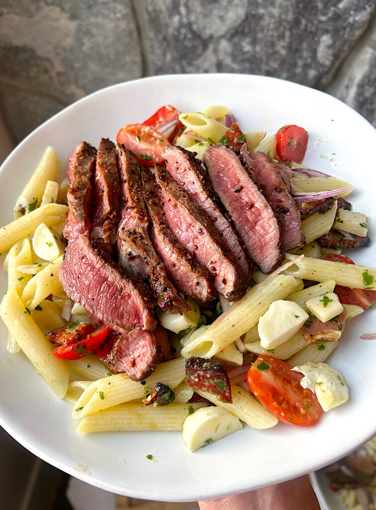 a white bowl of pasta salad topped with sliced steak