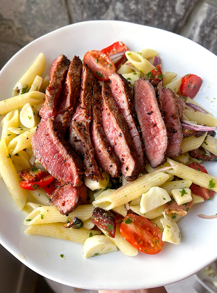 a white bow of pasta salad with tomatoes, mozzarella, steak and a basil dressing