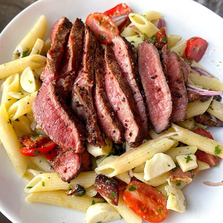 a white bow of pasta salad with tomatoes, mozzarella, steak and a basil dressing