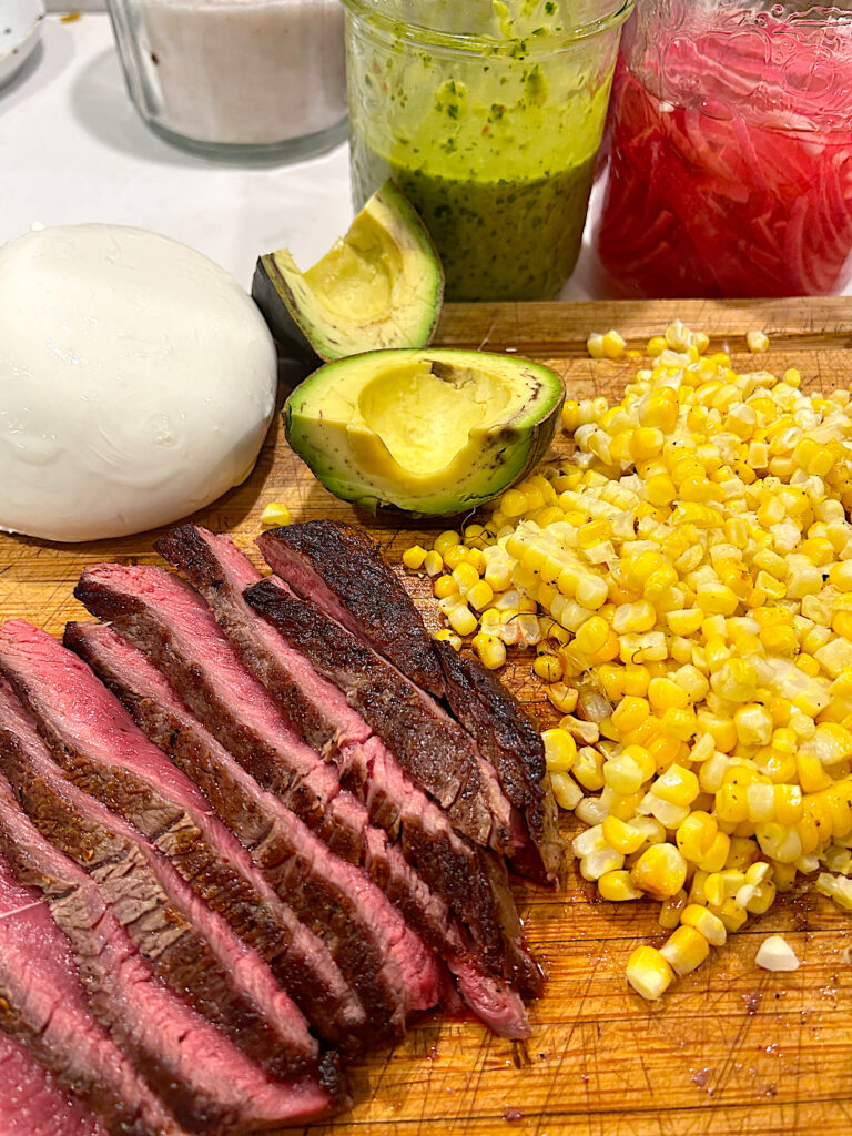 a wood board with sliced steak, corn, avocado and a ball of burrata cheese