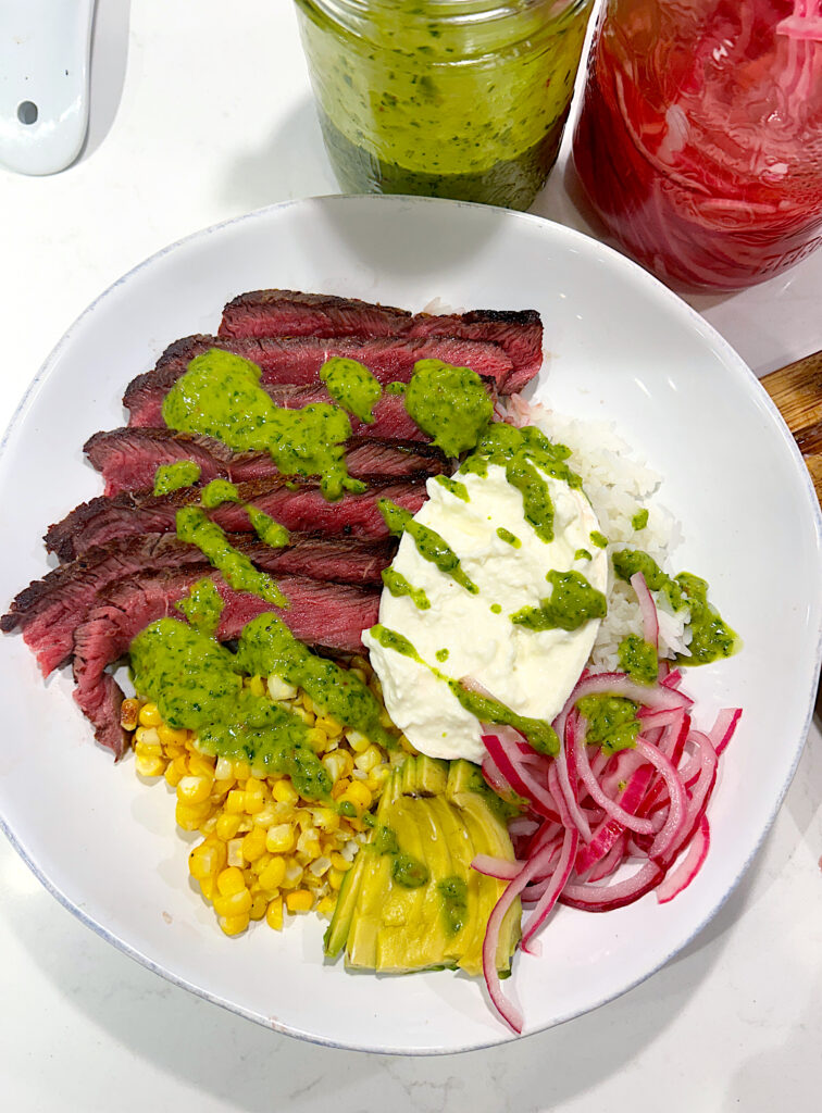 a rice bowl with sliced steak, burrata cheese, pickled onions and chimichurri sauce