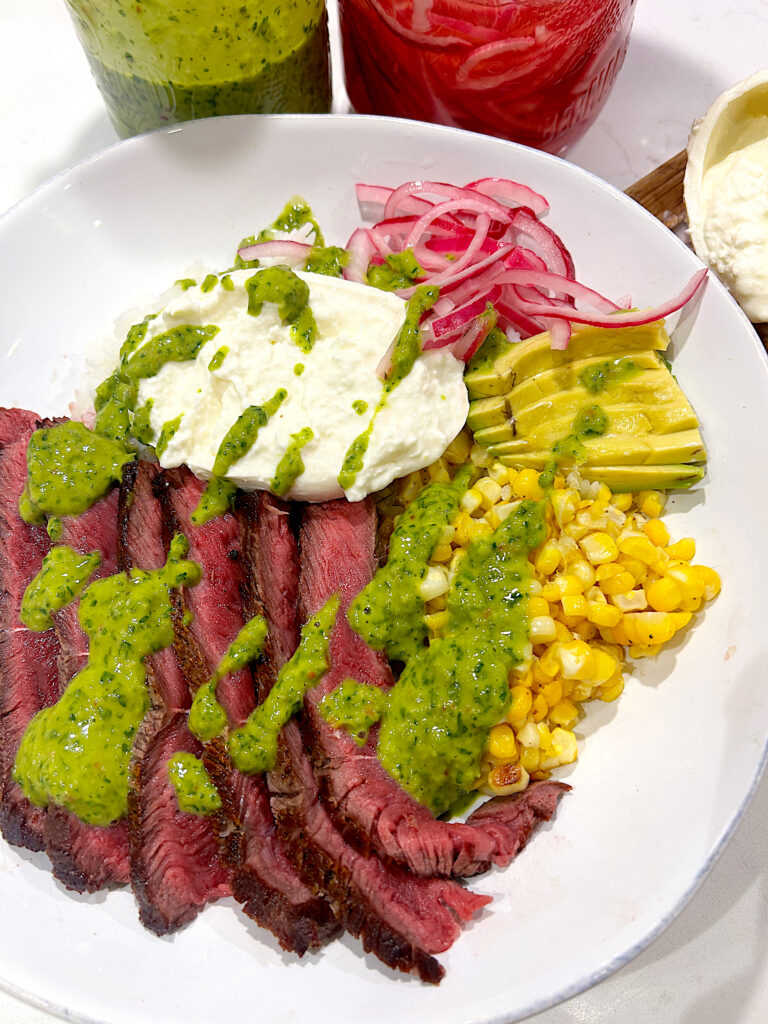 a steak bowl with burrata cheese, corn and chimichurri sauce