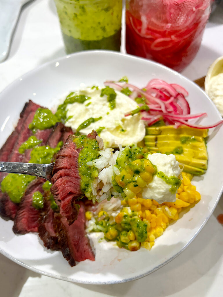a rice bowl with corn, pickled red onions, burrata cheese, steak and chimichurri