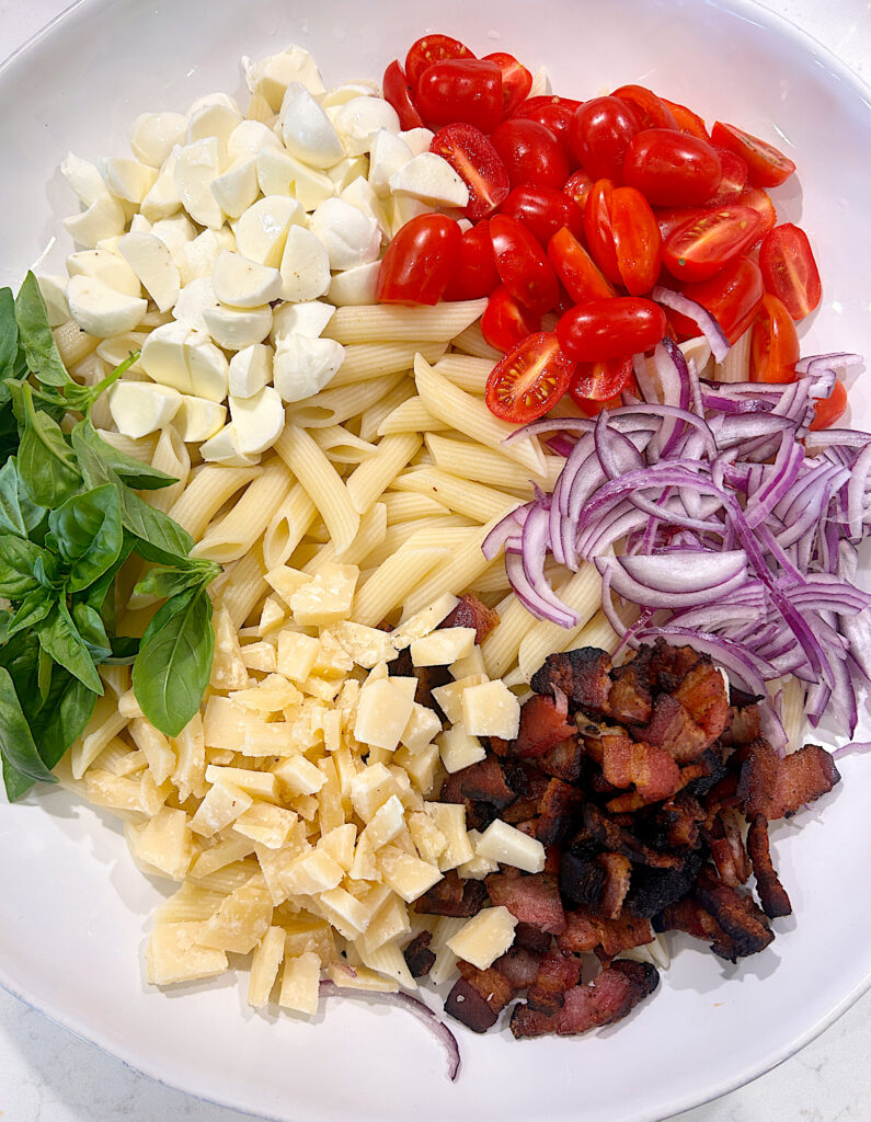 a large white bowl with pasta, tomatoes, cheese, bacon, red onion and basil