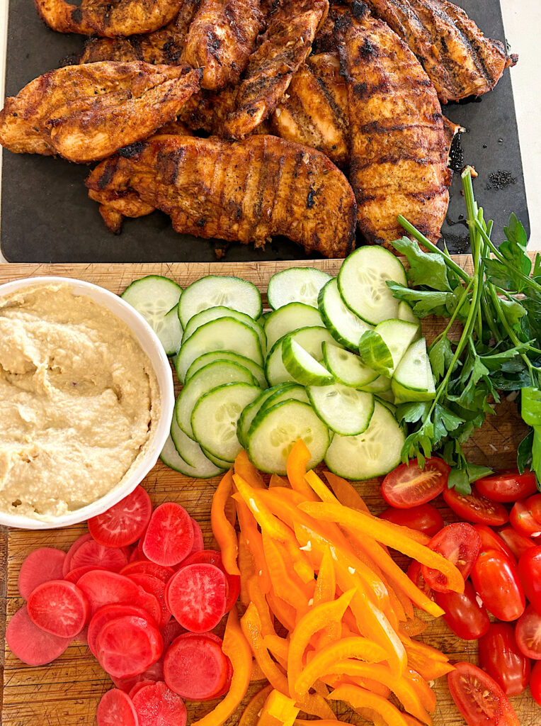 a wood board full of sliced vegetables, a bowl of hummus and grilled shawarma chicken