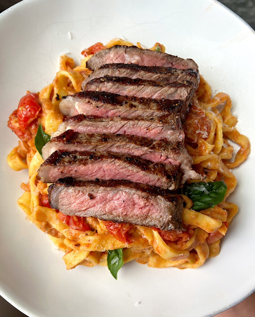 a white bowl of tomato and basil pasta with steak on top