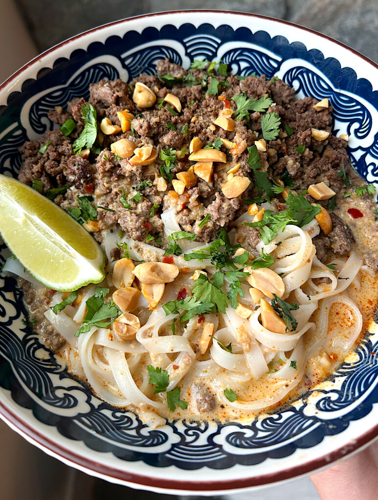 a blue and white bowl of creamy curry coconut broth with noodles, ground venison, peanuts and fresh lime