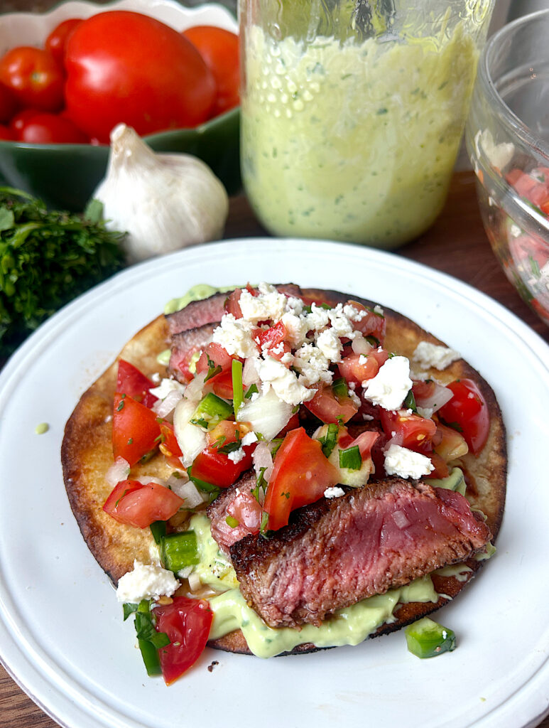 a white plate with a crispy tortilla topped with creamy avocado, sliced venison and salsa