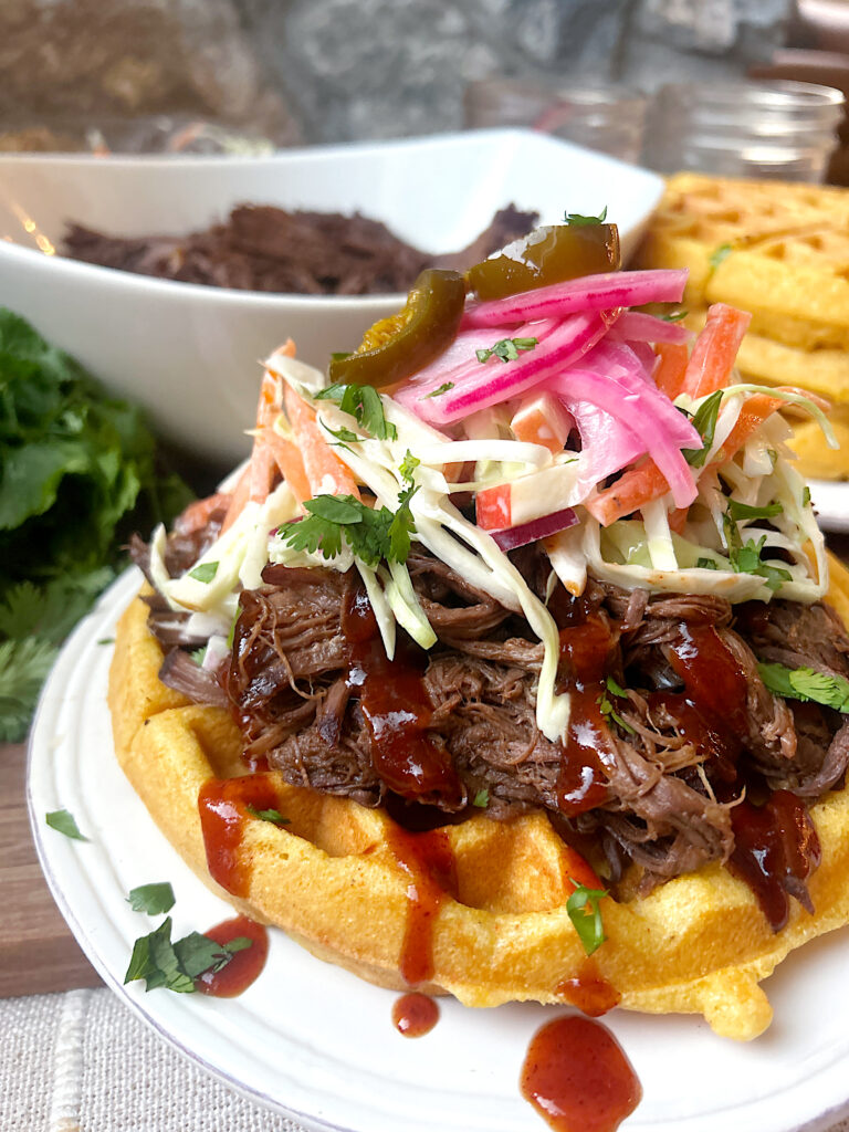a white plate with a cornbread waffle, topped with shredded bbq meat and coleslaw