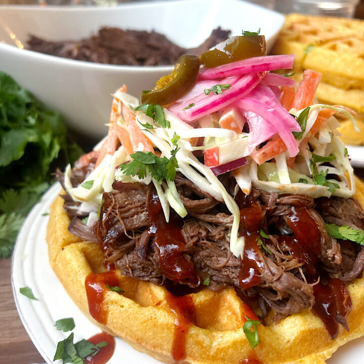 a white plate with a cornbread waffle, topped with shredded bbq meat and coleslaw