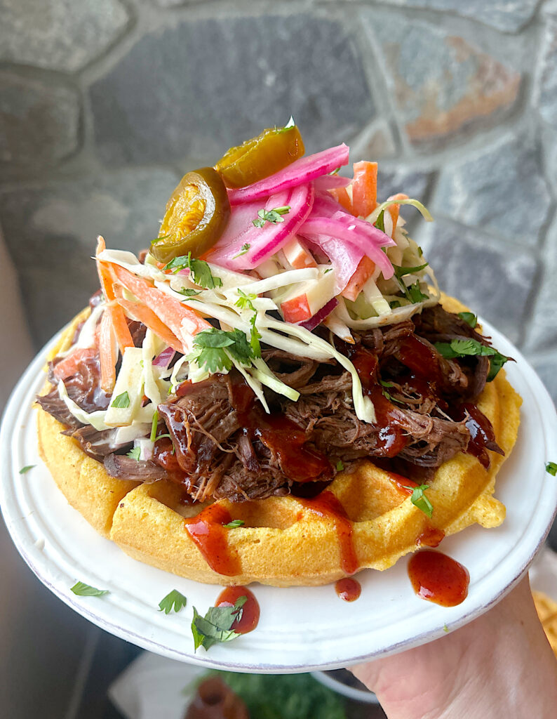 a white plate with a cornbread waffles topped with shredded bbq meat and coleslaw