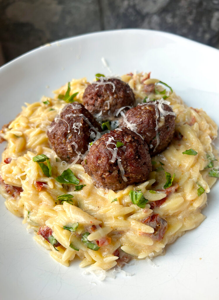 a white bowl of orzo pasta in a creamy white wine sauce with fresh basil and meatballs