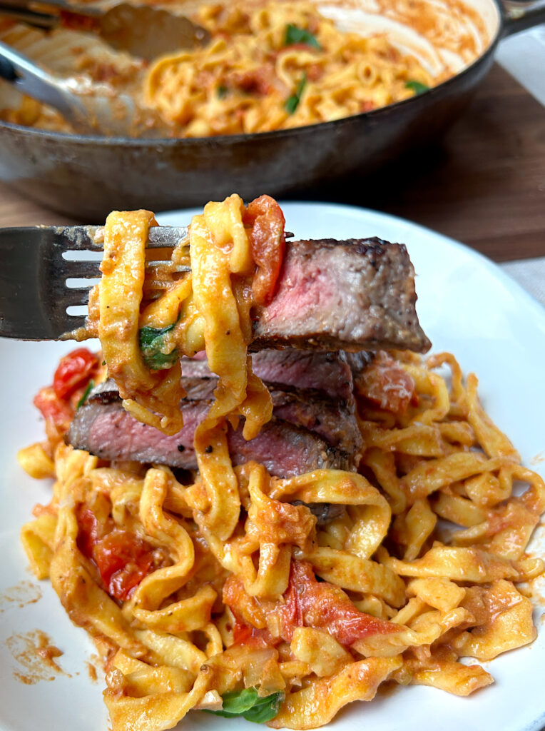 a fork full of cherry tomato pasta with a piece of steak