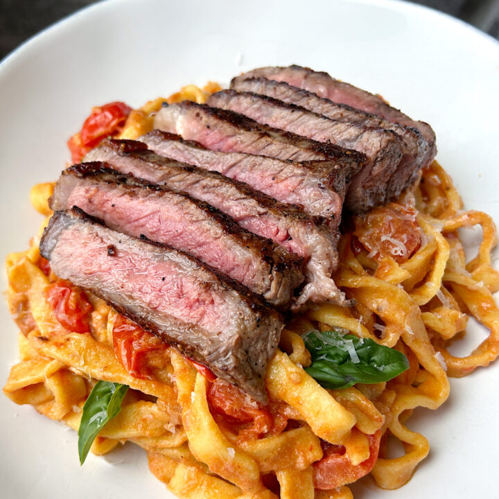 a white bowl of fettuccini noodles with a cherry tomato sauce and sliced steak