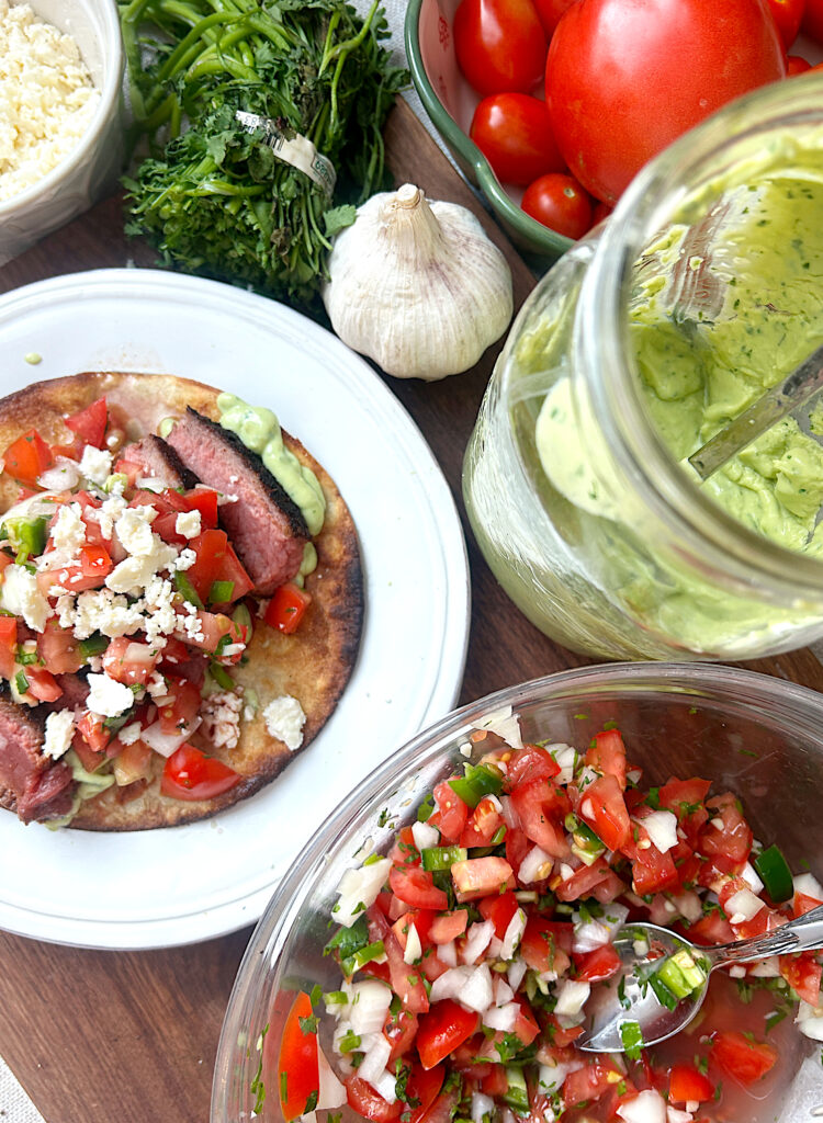 a white plate with a steak tostada, with a bowl of salsa, and a jar of avocado crema on the side
