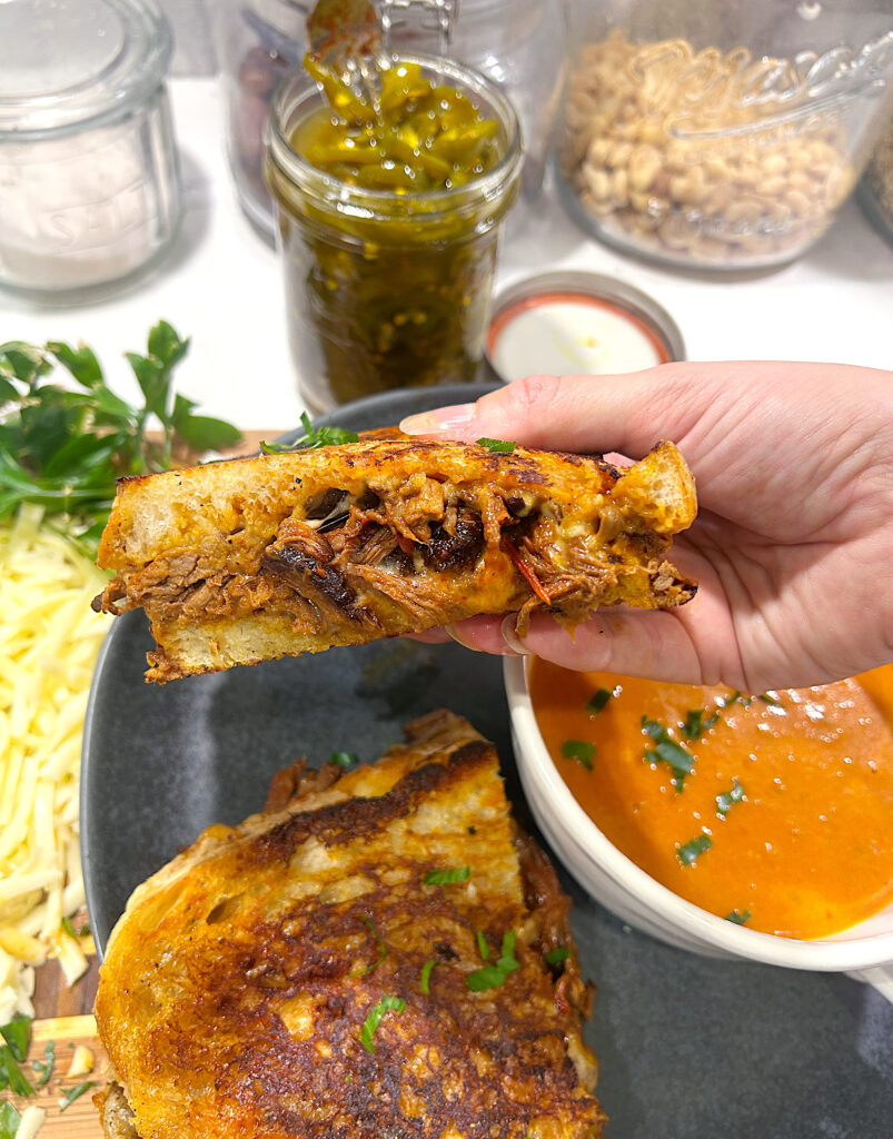 half of a grilled cheese with birria and a side of tomato soup