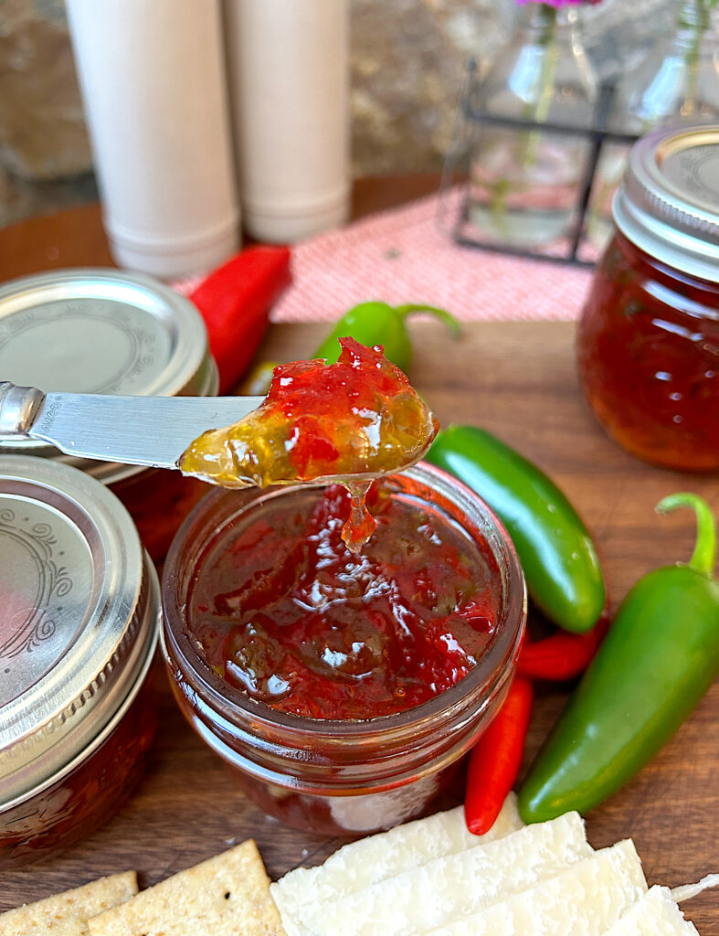 a jar of pepper jelly on a wood board with cheese and crackers