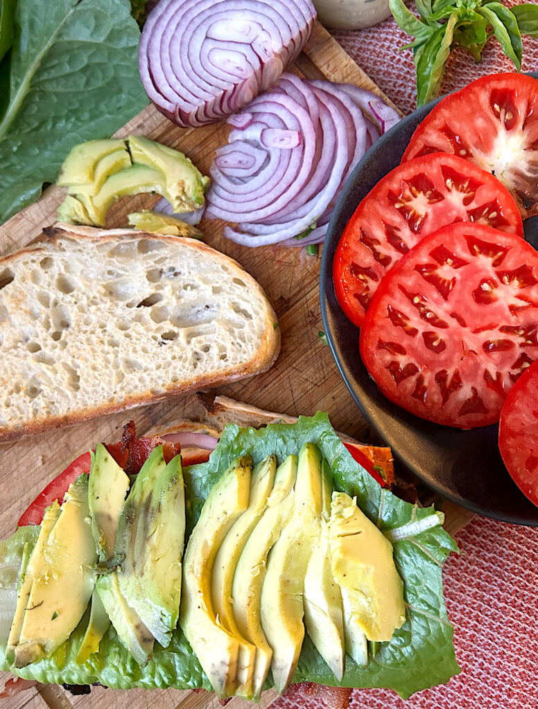 a plate of sliced tomatoes, red onion, avocado and sourdough bread on a wood board