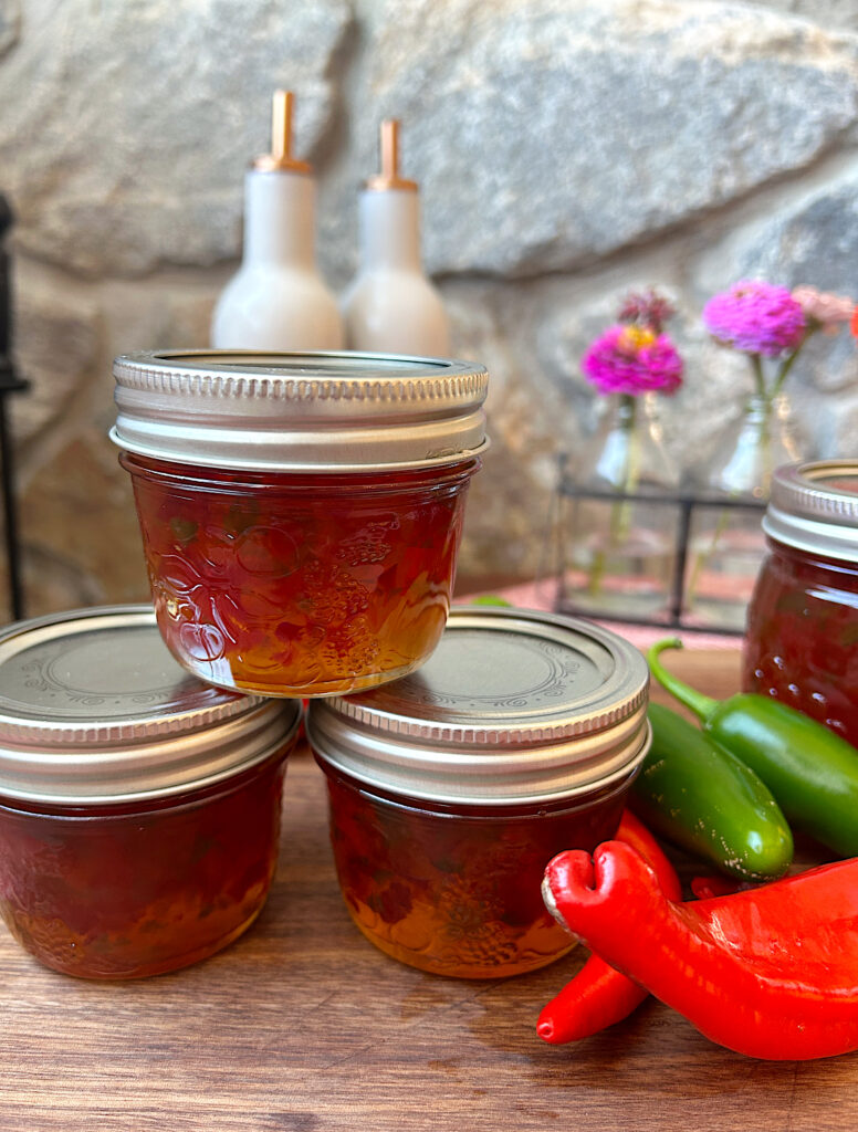 three small jars of jelly n a wood board with red and green peppers