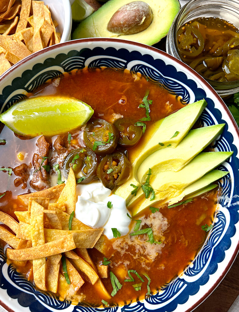 a blue and white bowl of tomato based soup topped with avocado, sour cream, lime and jalapenos