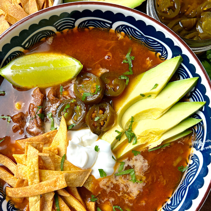 a blue and white bowl of tomato based soup topped with avocado, sour cream, lime and jalapenos