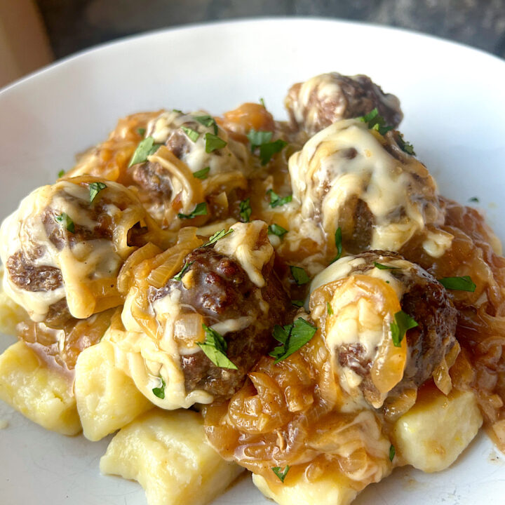 a white bowl of gnocchi topped with caramelized onions, meatballs and Swiss cheese
